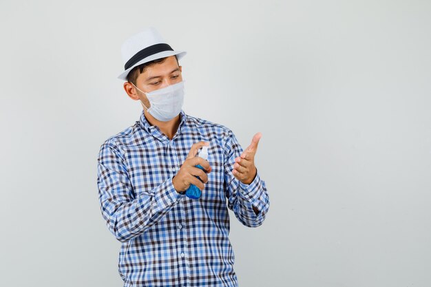 Junger Mann im karierten Hemd, im Hut, in der Maske, die Desinfektionsspray auf Hand aufträgt und vorsichtig schaut
