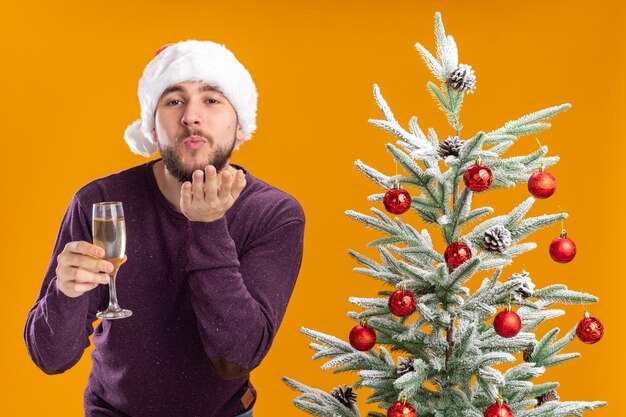 Junger Mann im lila Pullover und in der Weihnachtsmannmütze, die Glas Champagner neben Weihnachtsbaum über orange Hintergrund halten