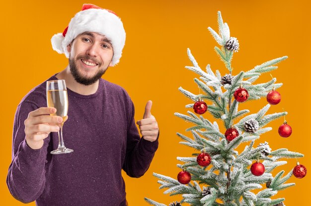 Junger Mann im lila Pullover und in der Weihnachtsmannmütze, die Glas Champagner neben Weihnachtsbaum über orange Hintergrund halten