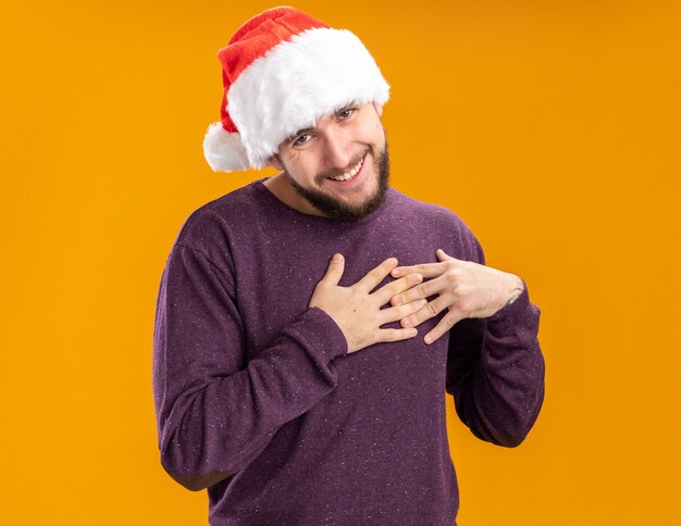 Junger Mann im lila Pullover und in der Weihnachtsmannmütze, die Kamera betrachtet, die Hände auf Brust hält, die dankbar sind, über orange Hintergrund zu stehen