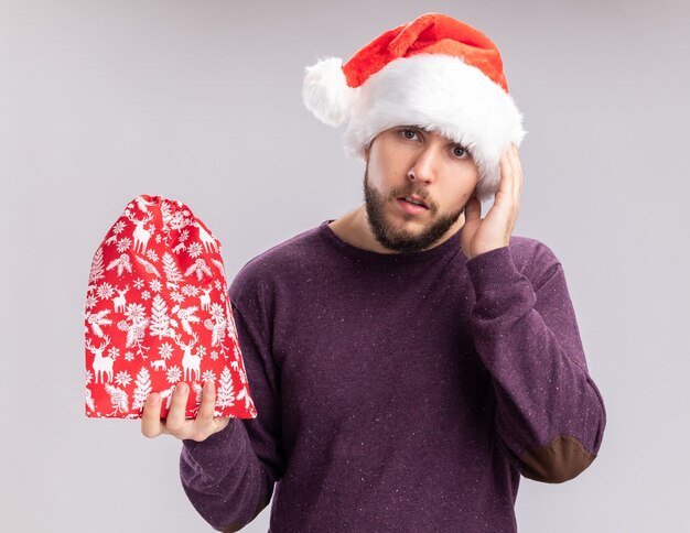 Junger Mann im lila Pullover und in der Weihnachtsmannmütze, die rote Tasche mit Geschenken hält, die Kamera verwirrt und sehr besorgt über weißem Hintergrund stehen