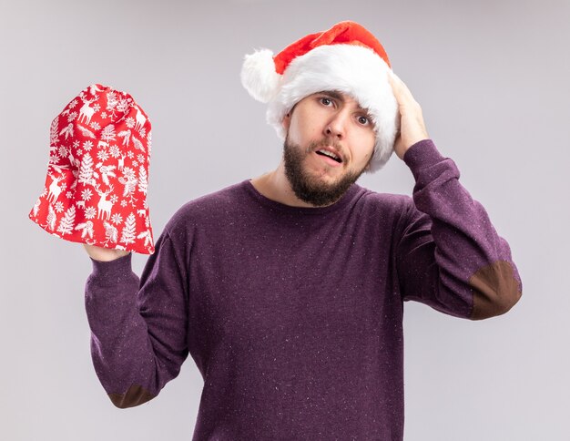 Junger Mann im lila Pullover und in der Weihnachtsmannmütze, die rote Tasche mit Geschenken hält, die mit Hand auf seinem Kopf für Fehler verwechselt suchen, der über weißer Wand steht