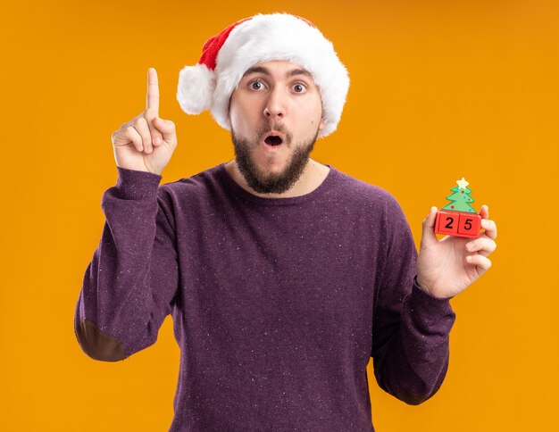 Junger Mann im lila Pullover und in der Weihnachtsmannmütze, die Spielzeugwürfel mit Nummer fünfundzwanzig überrascht zeigt Zeigefinger, der über orange Wand steht