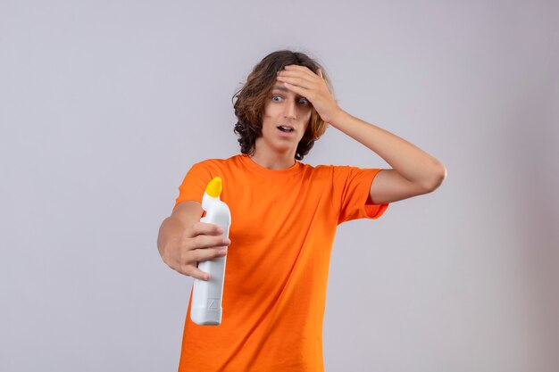 Junger Mann im orangefarbenen T-Shirt, das eine Flasche Reinigungsmittel hält, die es betrachtete, berührte den berührenden Kopf mit der Hand, die über weißem Hintergrund steht