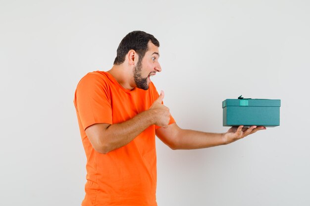 Junger Mann im orangefarbenen T-Shirt, das Geschenkbox mit Daumen nach oben hält und glücklich aussieht, Vorderansicht.