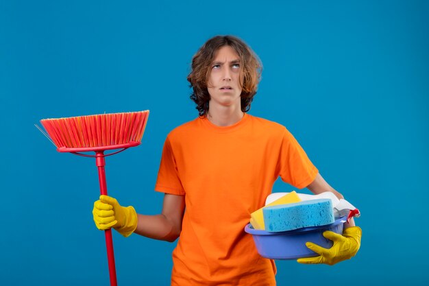 Junger Mann im orangefarbenen T-Shirt, das Gummihandschuhe hält, die Becken mit Reinigungswerkzeugen und Mopp halten, der verwirrt und ängstlich über blauem Hintergrund stehend schaut