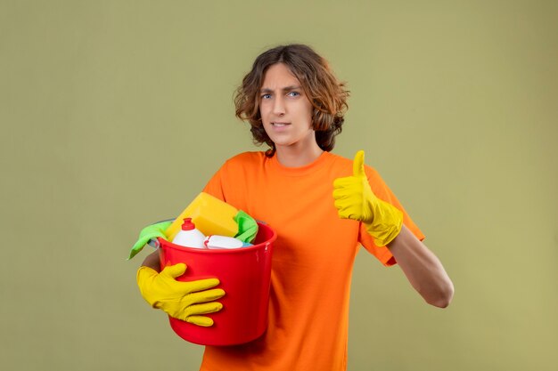 Junger Mann im orangefarbenen T-Shirt, das Gummihandschuhe hält, die Eimer mit Reinigungswerkzeugen betrachten, die Kamera betrachten, die fröhlich Daumen hoch zeigt, die über grünem Hintergrund stehen
