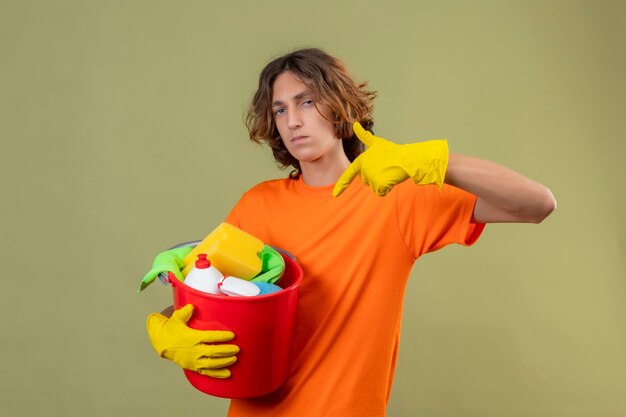 Junger Mann im orangefarbenen T-Shirt, das Gummihandschuhe hält, die Eimer mit Reinigungswerkzeugen halten, die mit dem Finger auf sie schauen, die t Kamera mit selbstbewusstem und ernstem Gesichtsausdruck stehen, der über gre steht