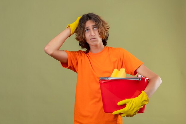 Junger Mann im orangefarbenen T-Shirt, das Gummihandschuhe hält, die Eimer mit Reinigungswerkzeugen halten, die mit Hand auf Kopf für Fehler suchen verwirrtes Denken stehen über grünem Hintergrund stehen