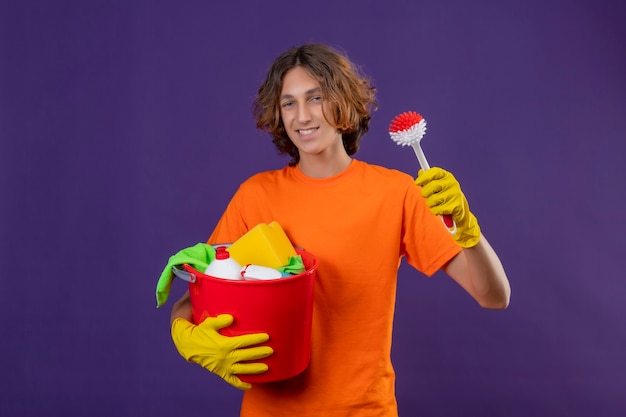 Junger Mann im orangefarbenen T-Shirt, das Gummihandschuhe hält, die Eimer mit Reinigungswerkzeugen und Schrubbbürste halten, die Kamera betrachten, die fröhlich über lila Hintergrund stehend lächelt