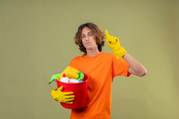 Junger Mann im orangefarbenen T-Shirt, das Gummihandschuhe hält, die Eimer mit Reinigungswerkzeugen zeigen, die große Idee haben, die zuversichtlich über grünem Hintergrund lächelnd steht