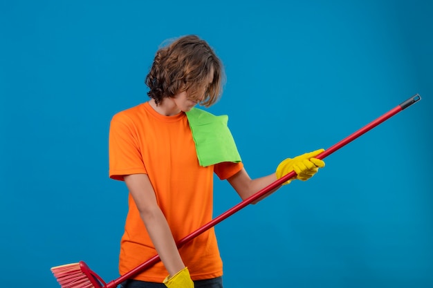 Junger Mann im orangefarbenen T-Shirt, das Gummihandschuhe hält, die Mopp halten, der als Gitarre verwendet, das Spaß hat, freudig stehend über blauem Hintergrund zu stehen