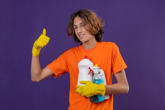 Junger Mann im orangefarbenen T-Shirt, das Gummihandschuhe hält, die Reinigungswerkzeuge halten, die Kamera mit dem selbstbewussten Lächeln betrachten, das Daumen oben steht über lila Hintergrund
