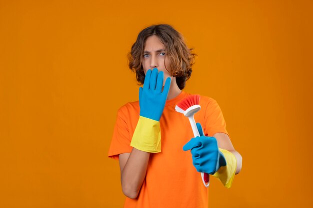 Junger Mann im orangefarbenen T-Shirt, das Gummihandschuhe hält, die Schrubbbürste suchen, die schockiert bedeckt Mund mit Hand steht über gelbem Hintergrund