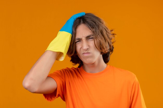 Junger Mann im orangefarbenen T-Shirt, das Gummihandschuhe mit Hand auf seinem Kopf für Fehler trägt, erinnert Fehler, vergaß schlechtes Gedächtniskonzept, das über gelbem Hintergrund steht