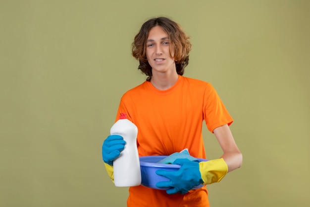 Junger Mann im orangefarbenen T-Shirt, das Gummihandschuhe trägt, die Becken mit Reinigungswerkzeugen und Flasche der Reinigungsmittel halten betrachten Kamera mit freundlichem Lächeln stehen über grünem Hintergrund