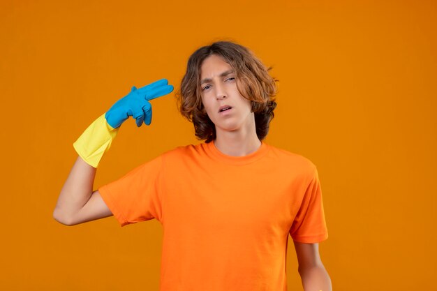 Junger Mann im orangefarbenen T-Shirt, das Gummihandschuhe trägt, die Fingerpistole oder Waffengeste nahe Tempel machen, die über gelbem Hintergrund stehen