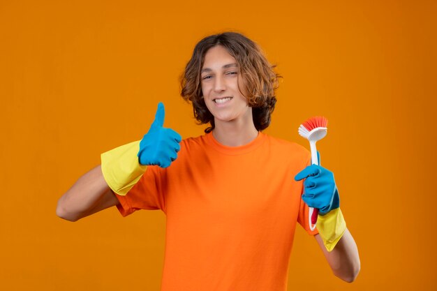 Junger Mann im orangefarbenen T-Shirt, das Gummihandschuhe trägt, die schrubbende Bürste lächelnd mit glücklichem Gesicht zeigen, das Daumen oben steht über gelbem Hintergrund