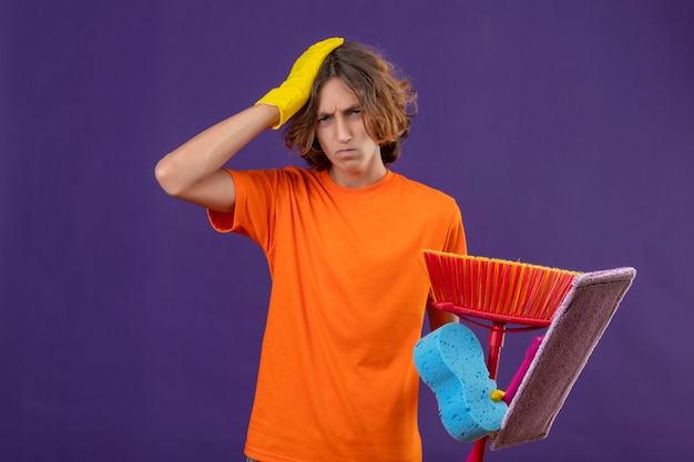 Junger mann im orangefarbenen t-shirt, das gummihandschuhe trägt, die schwamm und mops halten, die mit hand auf kopf für fehler stehen, der sehr besorgt über lila hintergrund schaut