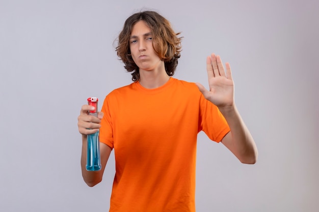 Junger Mann im orangefarbenen T-Shirt, das Reinigungsspray hält, das Stoppschild mit Handverteidigungsgeste betrachtet, die Kamera mit stirnrunzelndem Gesicht betrachtet, das über weißem Hintergrund steht