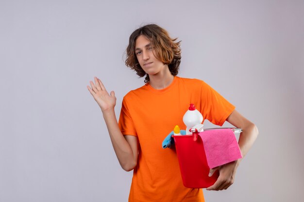 Junger Mann im orangefarbenen T-Shirt, der Eimer mit Reinigungswerkzeugen hält, die zuversichtlich lächelnd stehen mit erhöhtem Arm über weißem Hintergrund stehen