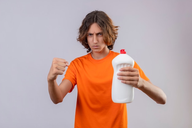 Junger Mann im orangefarbenen T-Shirt, der Flasche Reinigungsmittel hält, das Faust zur Kamera zeigt, die mit dem Stirnrunzeln drohend steht, das über weißem Hintergrund steht