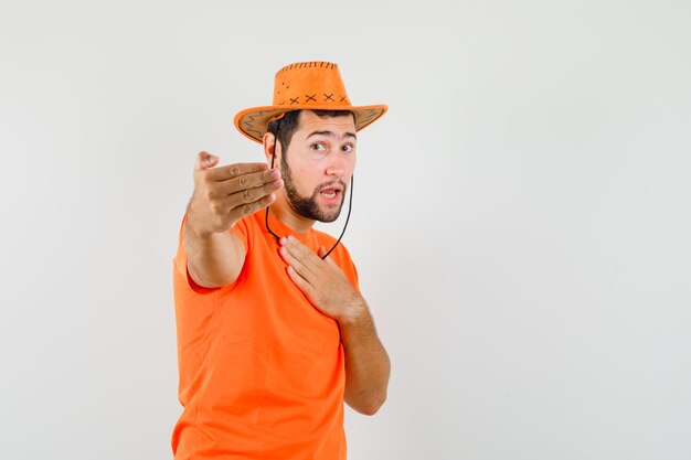 Junger Mann im orangefarbenen T-Shirt, Hut, der zum Kommen einlädt, Vorderansicht.