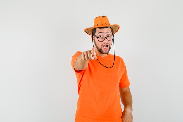 Junger Mann im orangefarbenen T-Shirt, Hut zeigt und sieht aufgeregt aus, Vorderansicht.