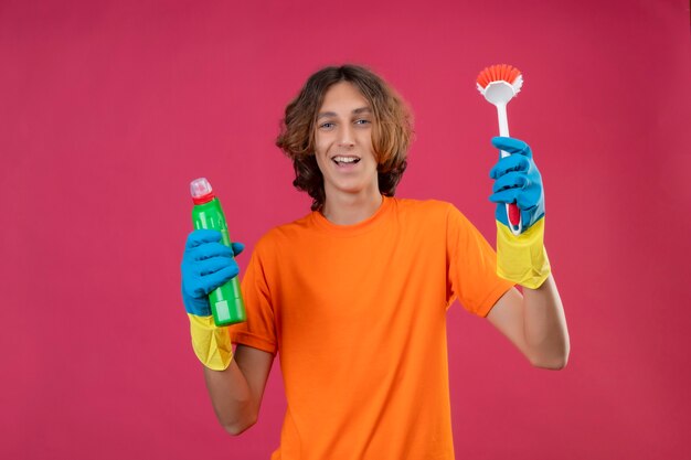 Junger Mann im orangefarbenen T-Shirt mit Gummihandschuhen, die eine Flasche Reinigungsmittel und eine Bürste mit Blick auf die Kamera halten, die fröhlich glücklich und positiv lächelnd über rosa Hintergrund lächelt