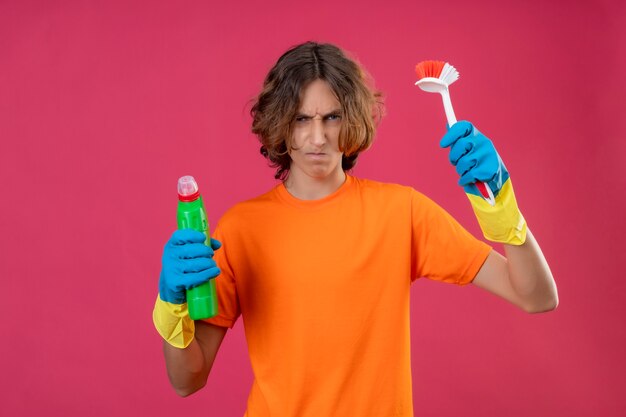 Junger Mann im orangefarbenen T-Shirt mit Gummihandschuhen, die eine Flasche Reinigungsmittel und eine Bürste mit Blick auf die Kamera halten, die mit dem stirnrunzelnden Gesicht über rosa Hintergrund unzufrieden ist