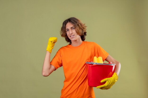 Junger Mann im orangefarbenen T-Shirt mit Gummihandschuhen, die einen Eimer mit Reinigungswerkzeugen halten, die die Faust ballen und sich über seinen Erfolg und Sieg freuen und über grünem Hintergrund glücklich stehen