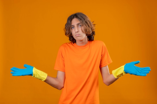 Junger Mann im orangefarbenen T-Shirt mit Gummihandschuhen zuckt mit den Schultern und sieht unsicher und verwirrt aus, ohne eine Antwort zu haben