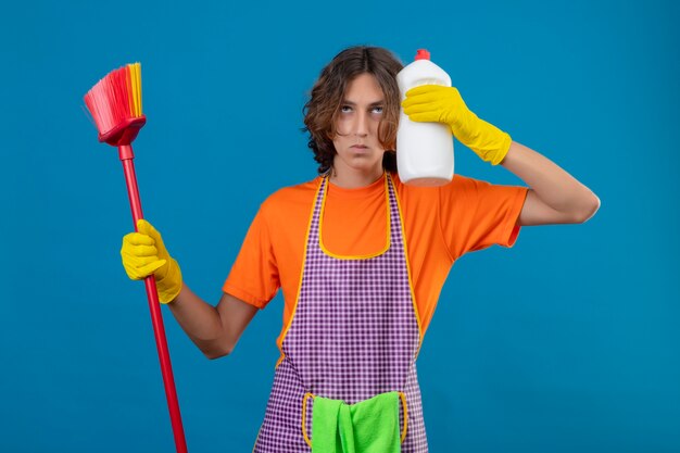 Junger Mann im orangefarbenen T-Shirt mit Schürze und Gummihandschuhen, die Mopp und Flasche Reinigungsmittel halten, die mit ernstem Gesicht suchen, das ängstlich über blauem Hintergrund steht