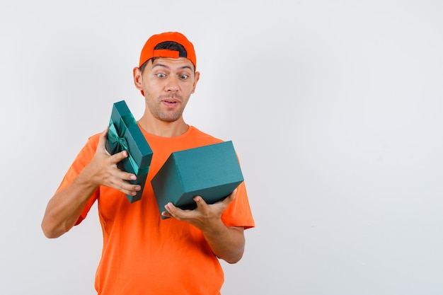 Junger Mann im orangefarbenen T-Shirt und in der Kappe, die in Geschenkbox schauen und erstaunt schauen