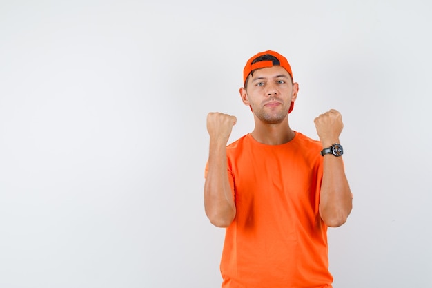 Junger Mann im orangefarbenen T-Shirt und in der Mütze, die erhobene Fäuste halten, geballt und zuversichtlich aussehend