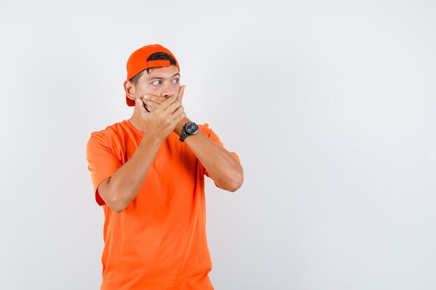 Junger Mann im orangefarbenen T-Shirt und in der Mütze, die Hände auf Mund halten und ängstlich schauen