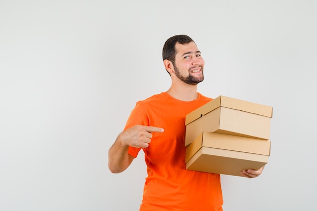 Junger Mann im orangefarbenen T-Shirt zeigt auf Kartons und sieht glücklich aus, Vorderansicht.