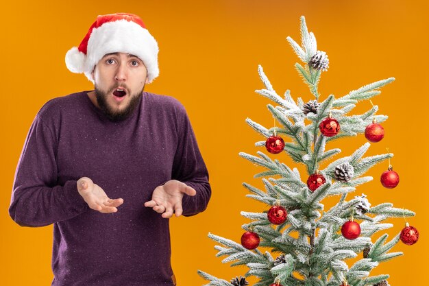 Junger Mann im purpurroten Pullover und in der Weihnachtsmannmütze, die Kamera betrachtet, verwirrt mit Armen, die in der Empörung angehoben werden, die neben Weihnachtsbaum über orange Hintergrund steht