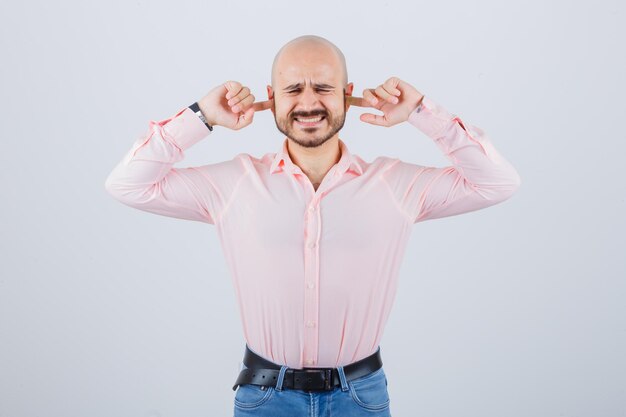 Junger Mann im rosa Hemd, Jeans, die Ohren mit den Fingern verstopfen, Vorderansicht.