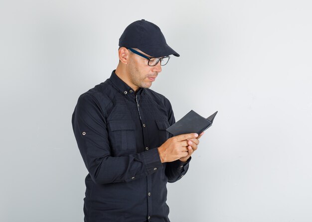 Junger Mann im schwarzen Hemd mit Kappe, Brille, die durch Notizbuch schaut
