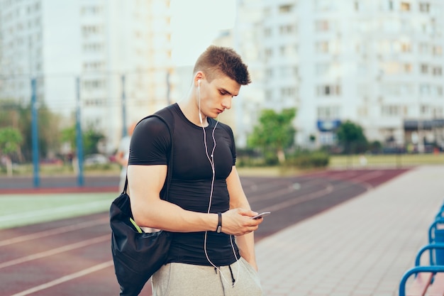 Junger Mann im Sportschwarzen T-Shirt steht am Morgen auf Stadt- und Stadionhintergrund. Er hört Musik über Kopfhörer, hält eine Sporttasche in der Hand und tippt am Telefon.