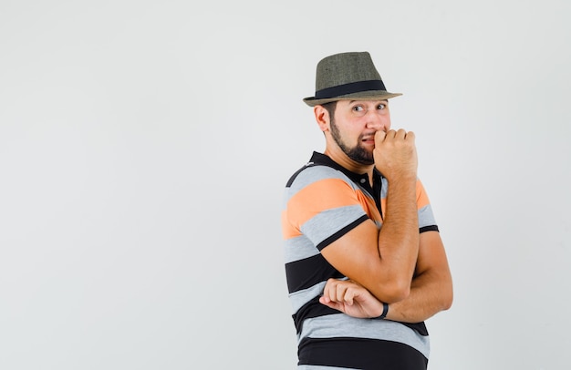 Junger Mann im T-Shirt, Hut, der in denkender Haltung steht und unruhig schaut, Vorderansicht.