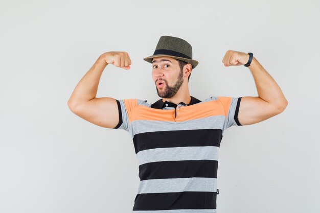 Junger Mann im T-Shirt, Hut zeigt seine Muskeln und sieht kraftvoll aus, Vorderansicht.