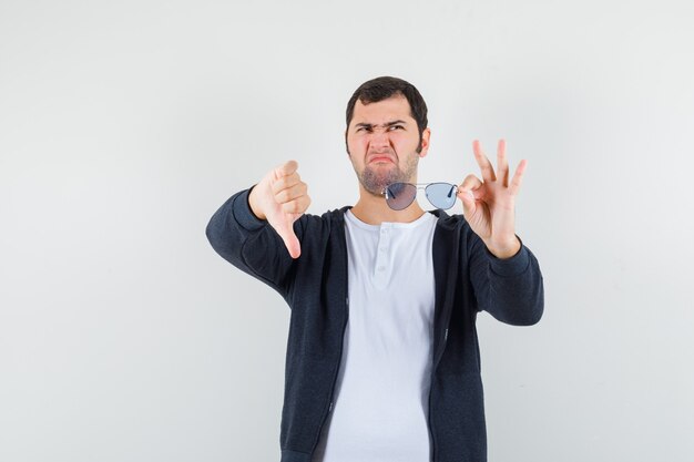 Junger Mann im T-Shirt, Jacke, die Brille hält, Daumen nach unten zeigt und Unzufriedenheit schaut, Vorderansicht.