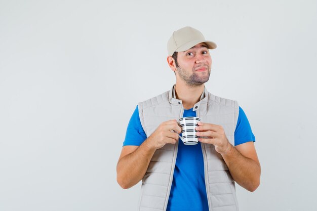 Junger Mann im T-Shirt, Jacke, die Tasse des Getränks hält und lustig schaut, Vorderansicht.