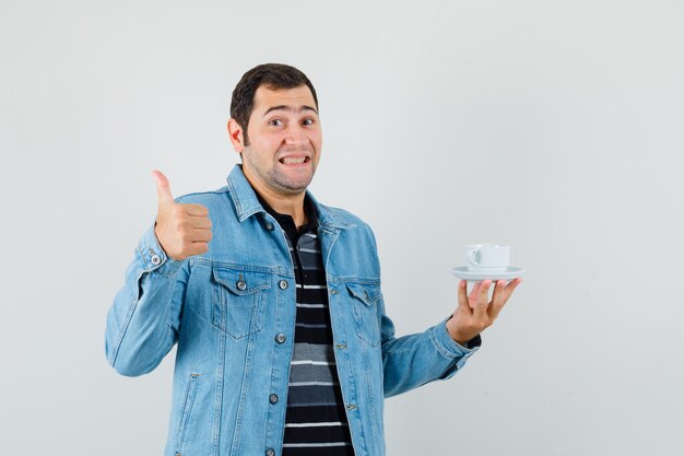 Junger Mann im T-Shirt, Jacke, die Tasse Tee hält, zeigt Daumen hoch und sieht glücklich aus