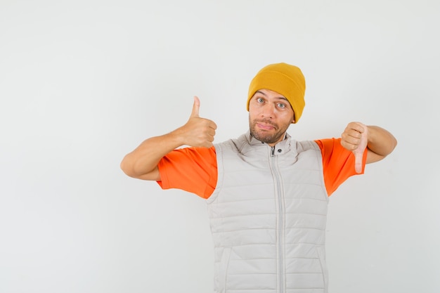 Junger Mann im T-Shirt, Jacke, Hut, der Daumen oben und unten zeigt, Vorderansicht.