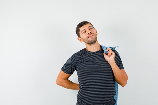 Junger Mann im T-Shirt posierend, während Jacke auf seinem Rücken haltend und niedlich aussehend