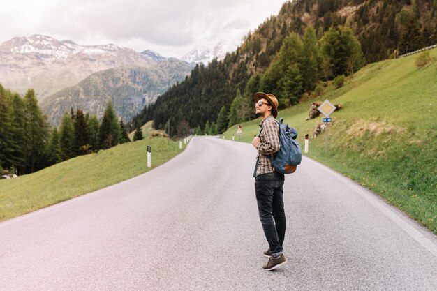 Junger Mann im trendigen Hut, der in der Mitte der Autobahn steht und wegschaut und frische Luft nahe italienischen Alpen genießt