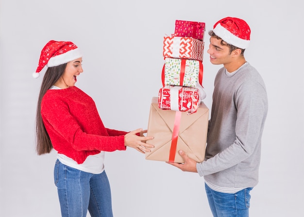 Junger Mann im Weihnachtshut mit Haufen von Geschenken nähert sich überraschter Frau